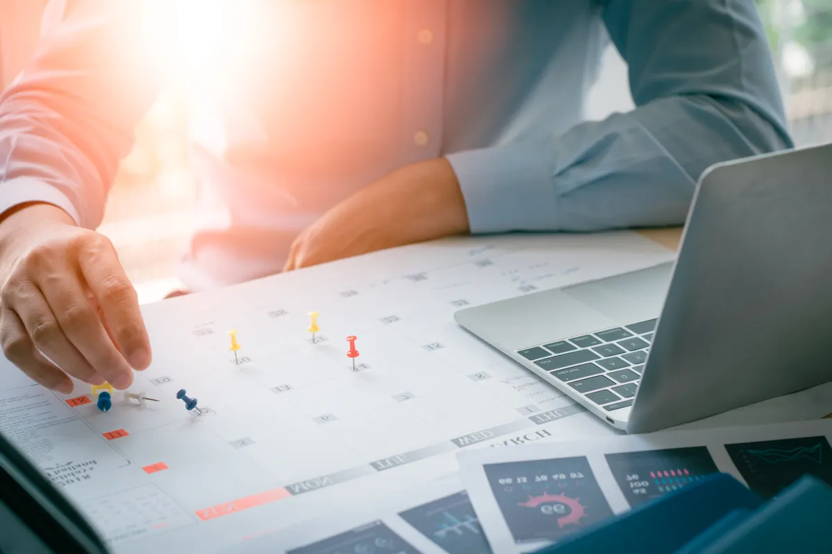 Pessoa marcando datas em um calendário de planejamento ao lado de um notebook.